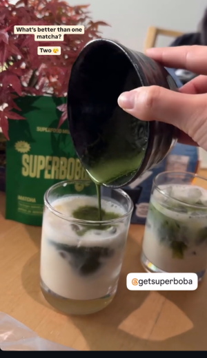 Hand pouring matcha into a glass of milk with a plant in the background.