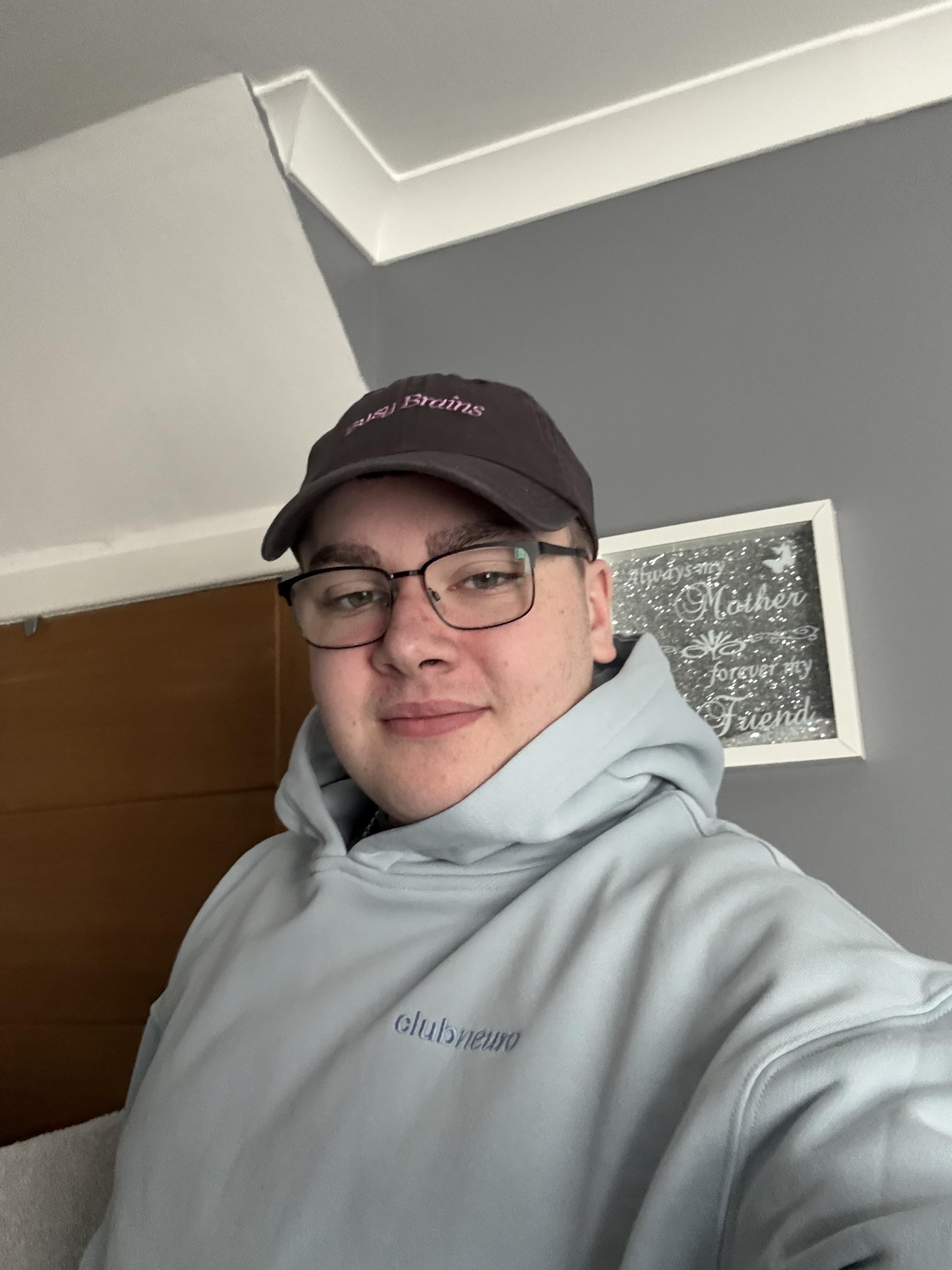 A person wearing glasses, a baseball cap, and a light blue hoodie takes a selfie in a room.