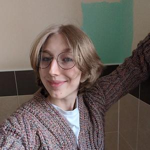 A young person with shoulder-length blonde hair and glasses, smiling while wearing a chunky knit cardigan.