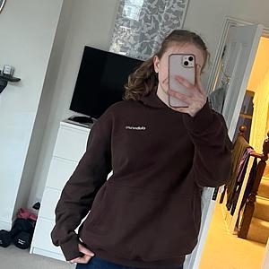 A young woman in a dark brown hoodie takes a mirror selfie with her smartphone in her room.