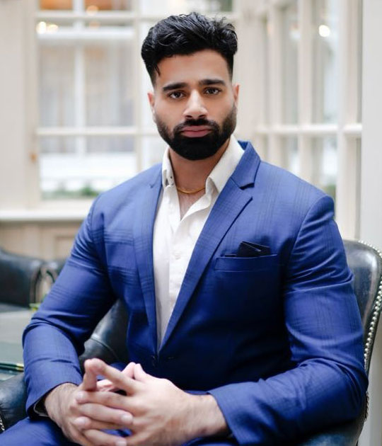 Man in blue suit seated indoors with hands clasped.