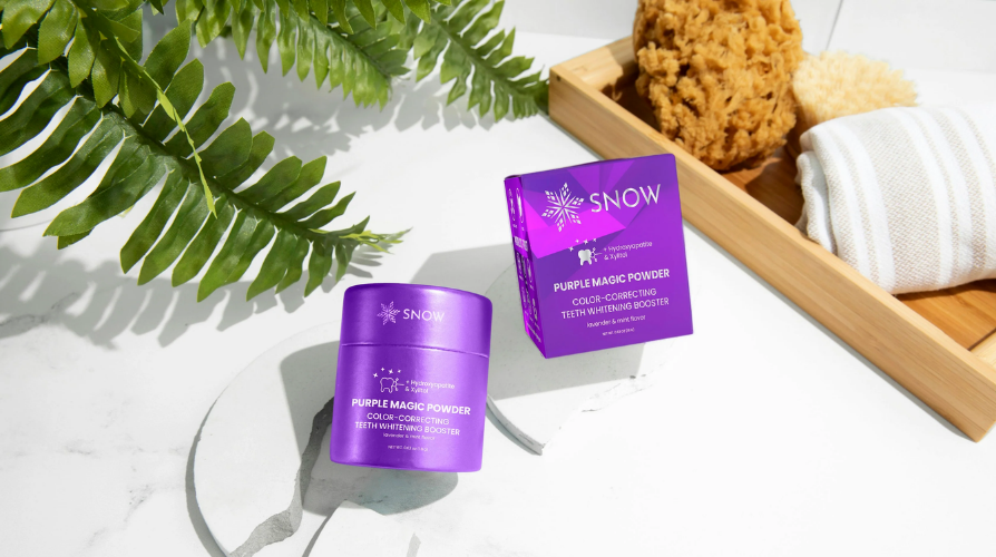 Purple tooth whitening powder on a bathroom counter with a plant and bath items in the background.