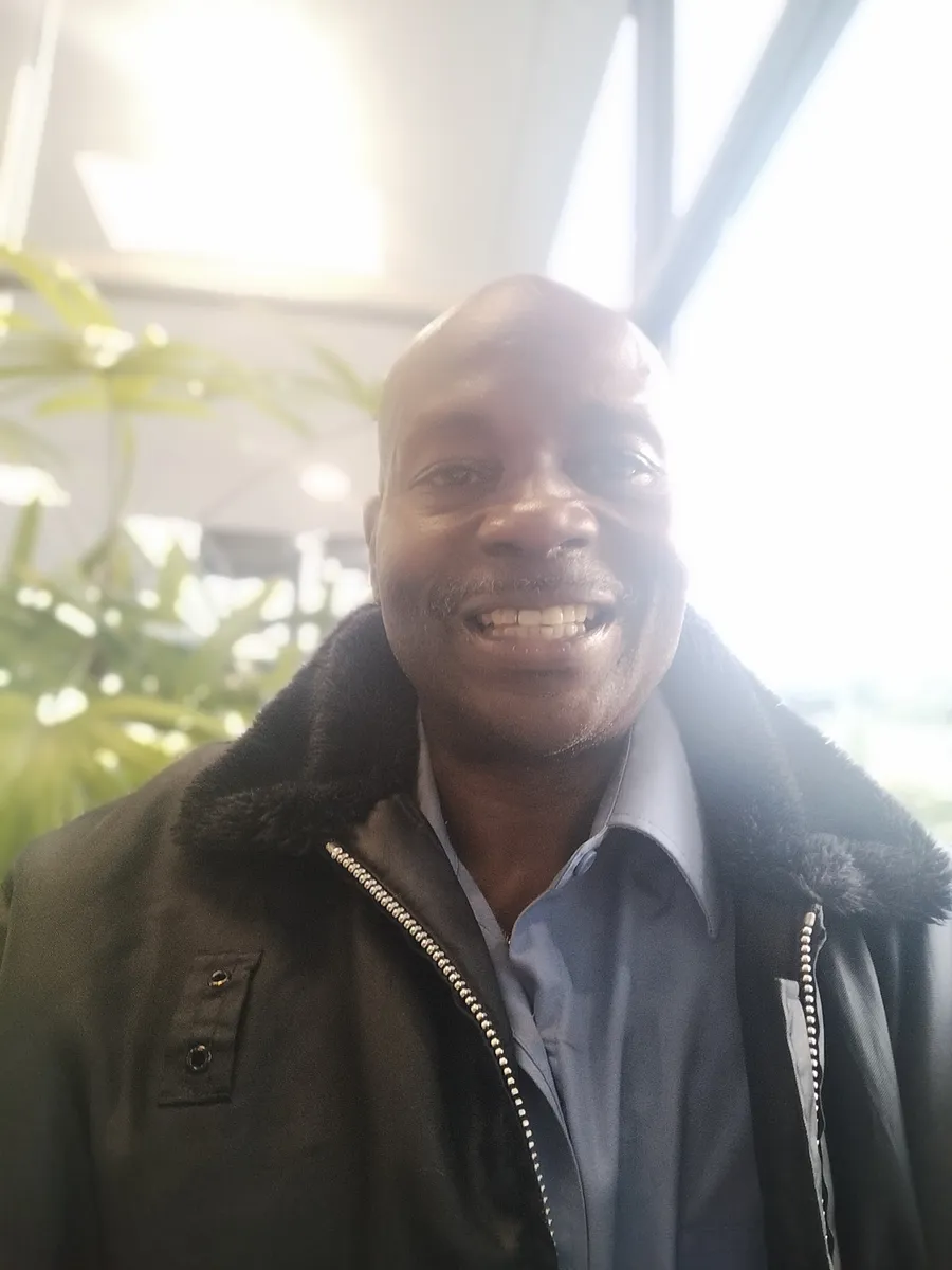 Smiling person in a jacket and shirt indoors with plants in the background.