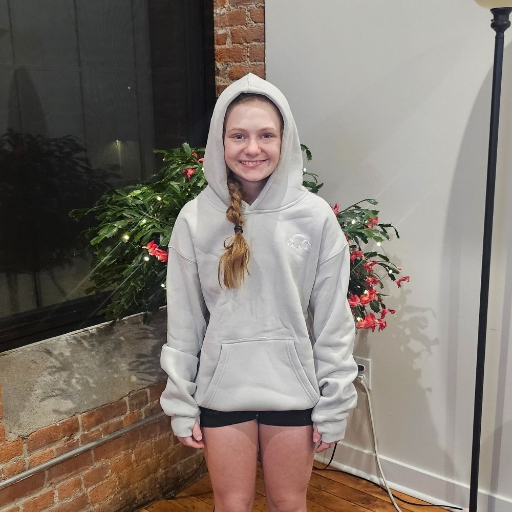 Person in a gray hoodie standing indoors with plants and a brick wall background.