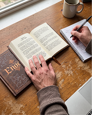 A person at a wooden desk writing in a notebook with an open book and a coffee mug.