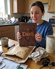 A woman sits at a kitchen table, excitedly holding up a copy of 'The Book of Enoch.'