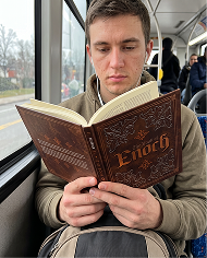 A young man with earphones sits on a bus, reading a book with 'Enoch' on the cover.