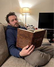 A man with a beard sits on a couch, reading a book titled 'Enoch'.