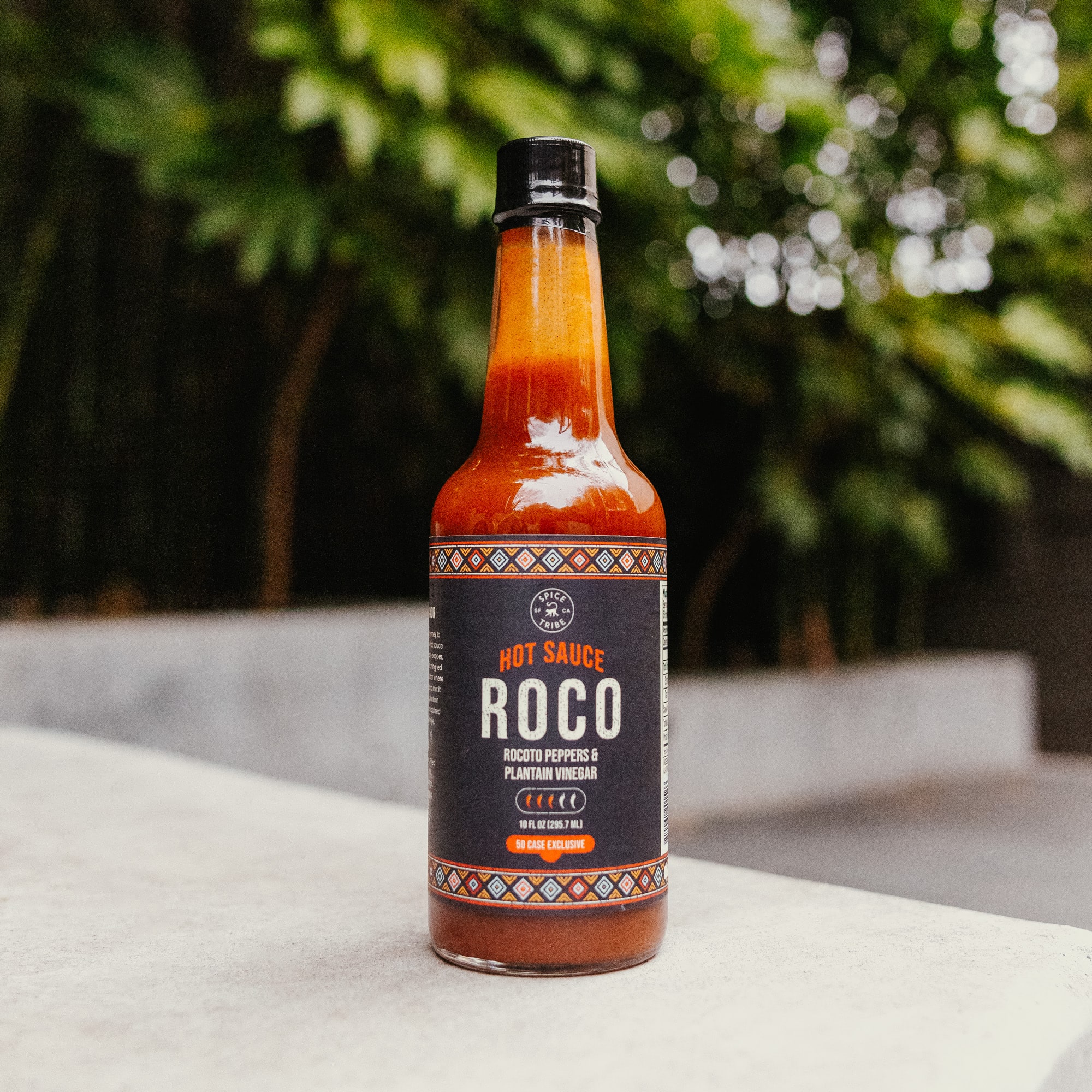 Bottle of Roco hot sauce on a concrete surface with greenery in the background.
