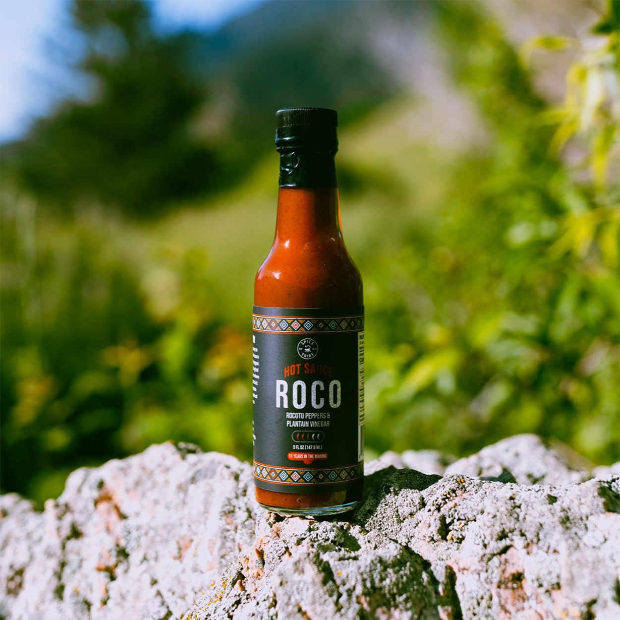 A bottle of Roco hot sauce on a rock with greenery in the background.