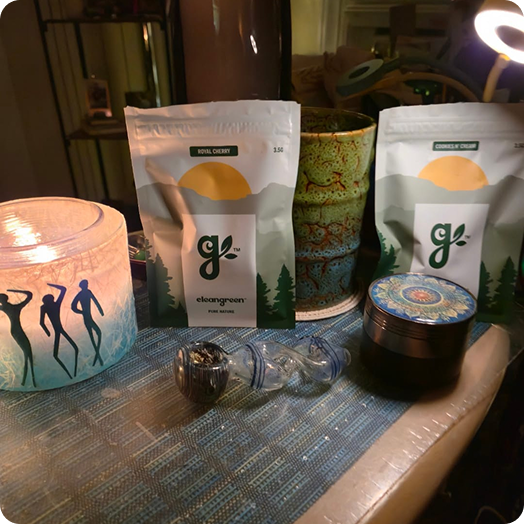 Two pouches of cannabis flower, a glass pipe, a grinder, and a lit candle on a tabletop.
