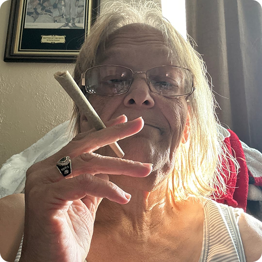 A close-up of an older woman with glasses holding a large, hand-rolled cigarette between her fingers.