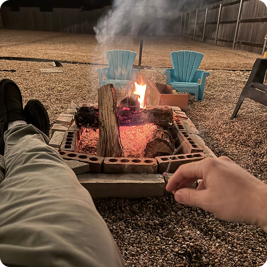 A person's legs and hand are visible in front of a brick fire pit at night.