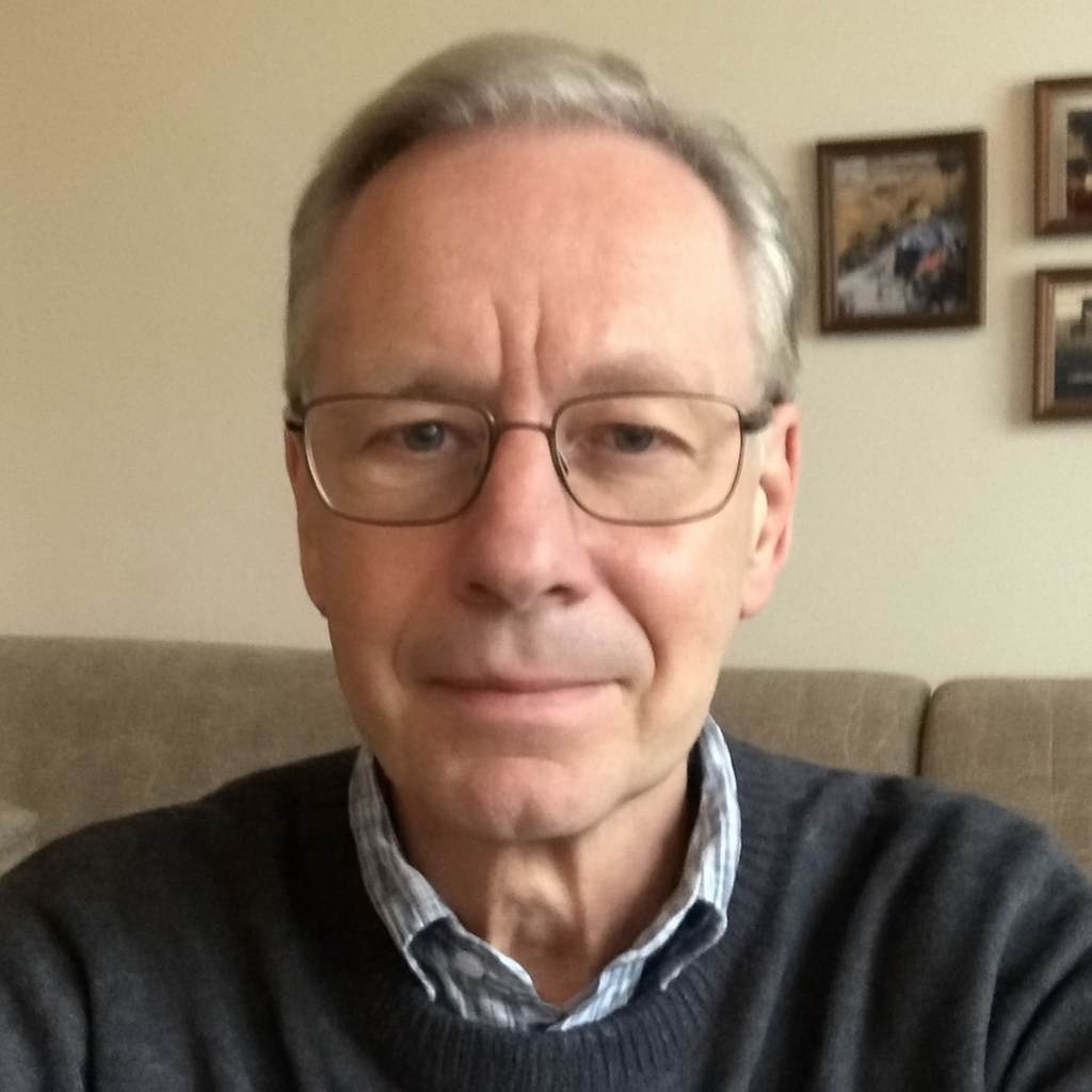 A person with glasses sitting on a couch, with framed pictures on the wall behind.
