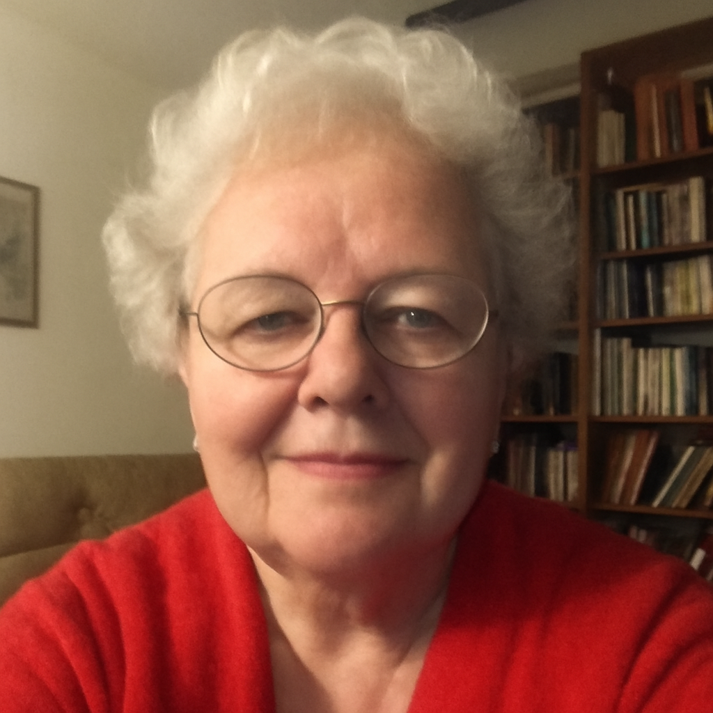 Elderly person with glasses and white hair, wearing a red sweater, in a room with bookshelves.