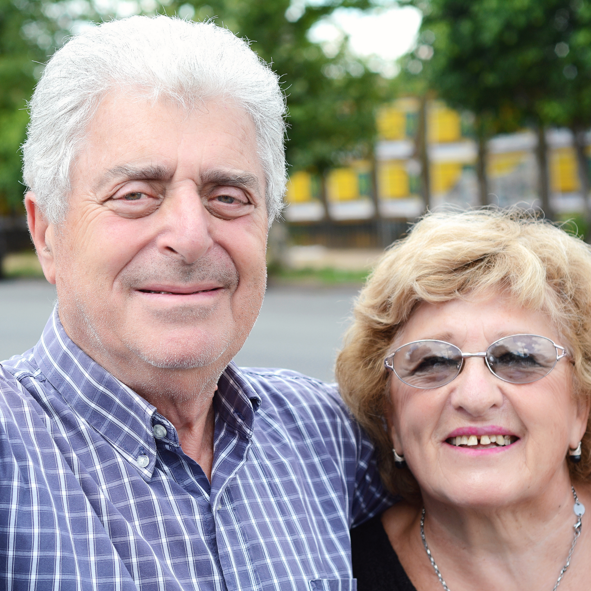Elderly couple smiling outdoors with trees in the background.