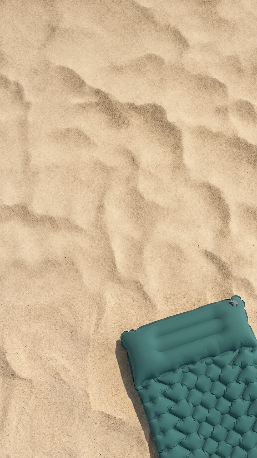 A top-down view of a teal-colored inflatable sleeping pad resting on sand.