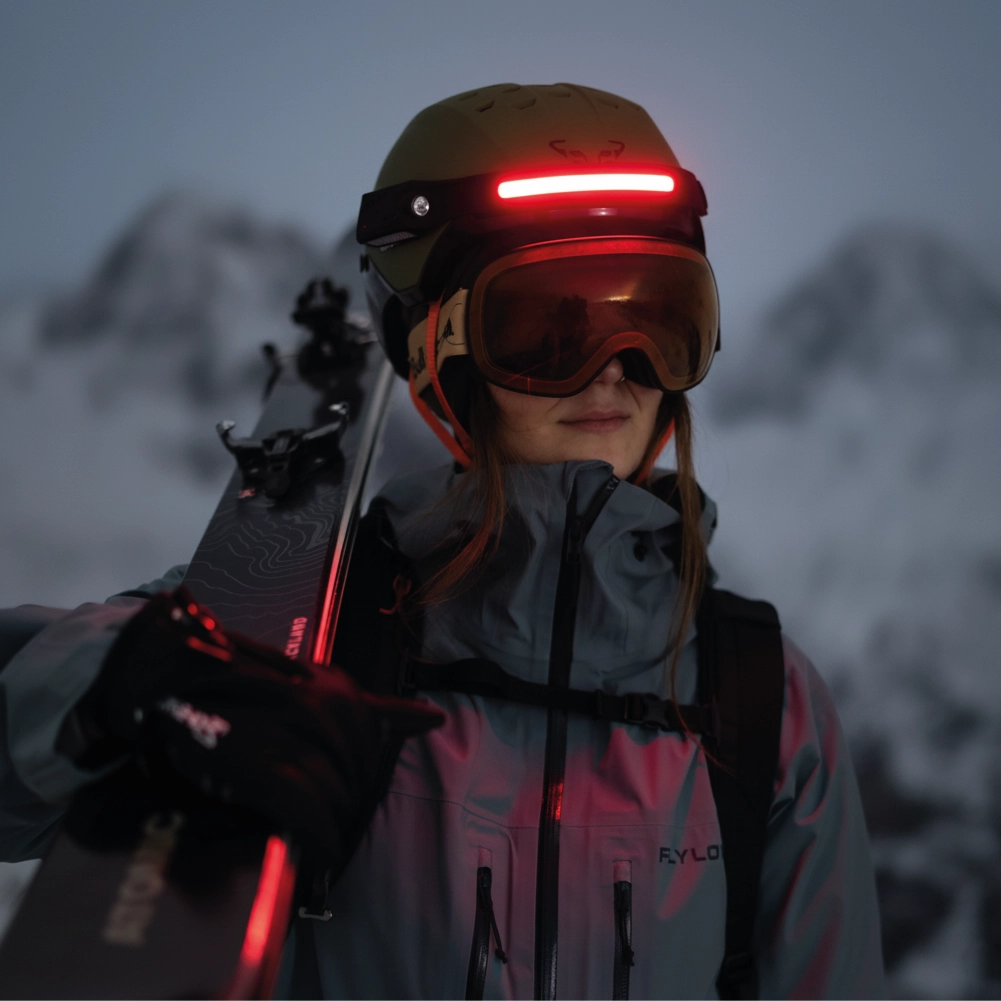 Person in ski gear with helmet and goggles, holding skis against a snowy mountainous background.