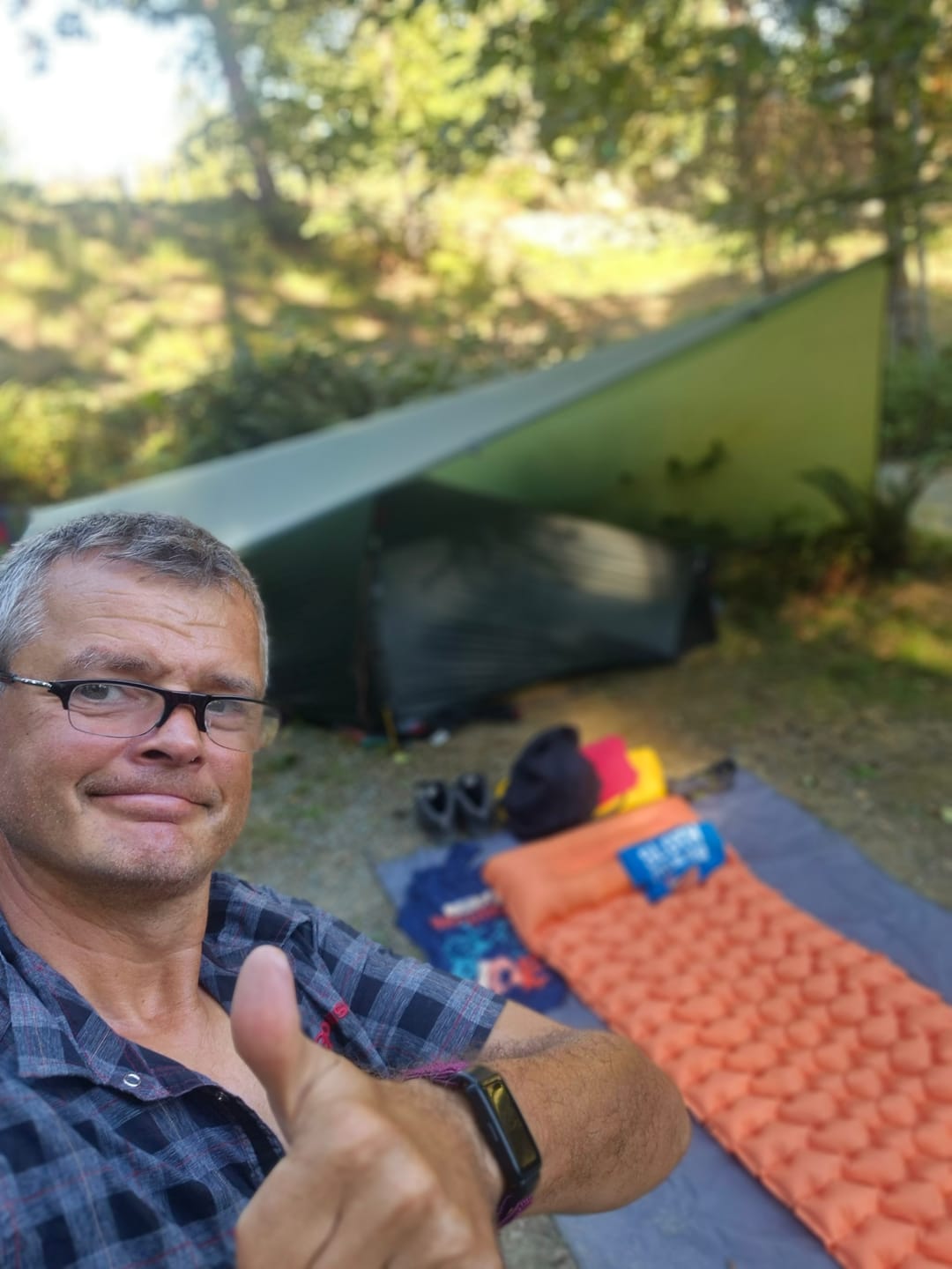 Man giving thumbs up in front of camping setup with sleeping mat and tarp.