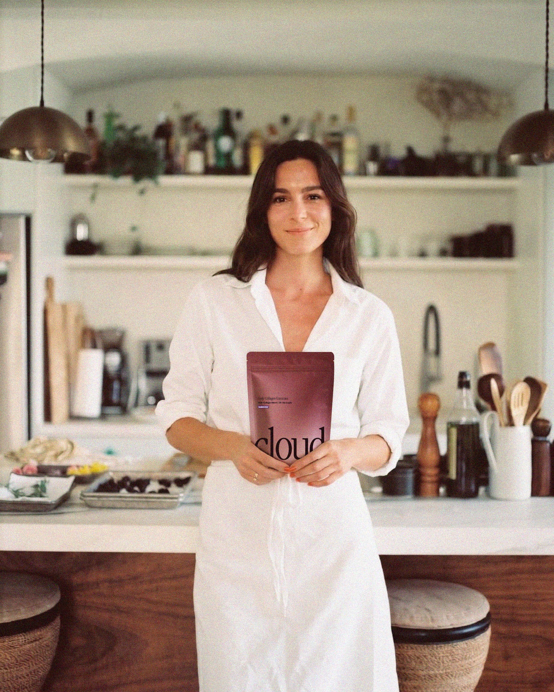 A woman in a white outfit stands in a kitchen holding a purple pouch labeled 'cloud.'