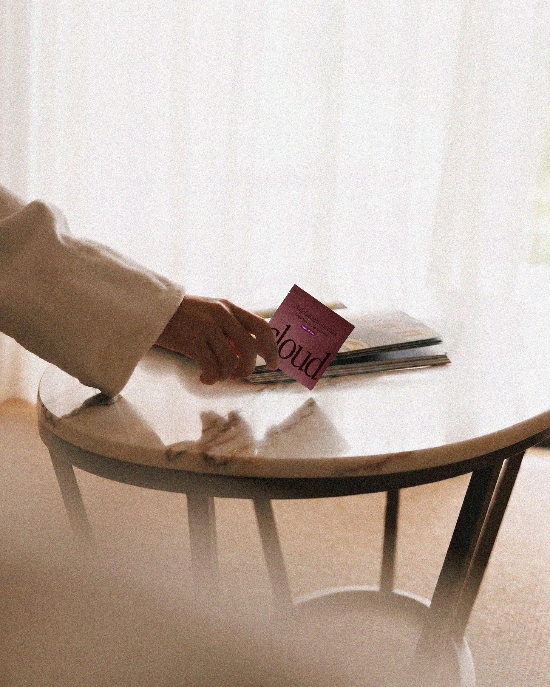 A hand in a white sleeve holds a small purple packet over a marble table with magazines.
