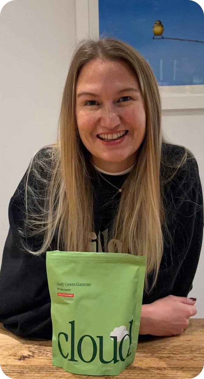 A smiling blonde woman in a black sweatshirt sits at a table with a green pouch in front.