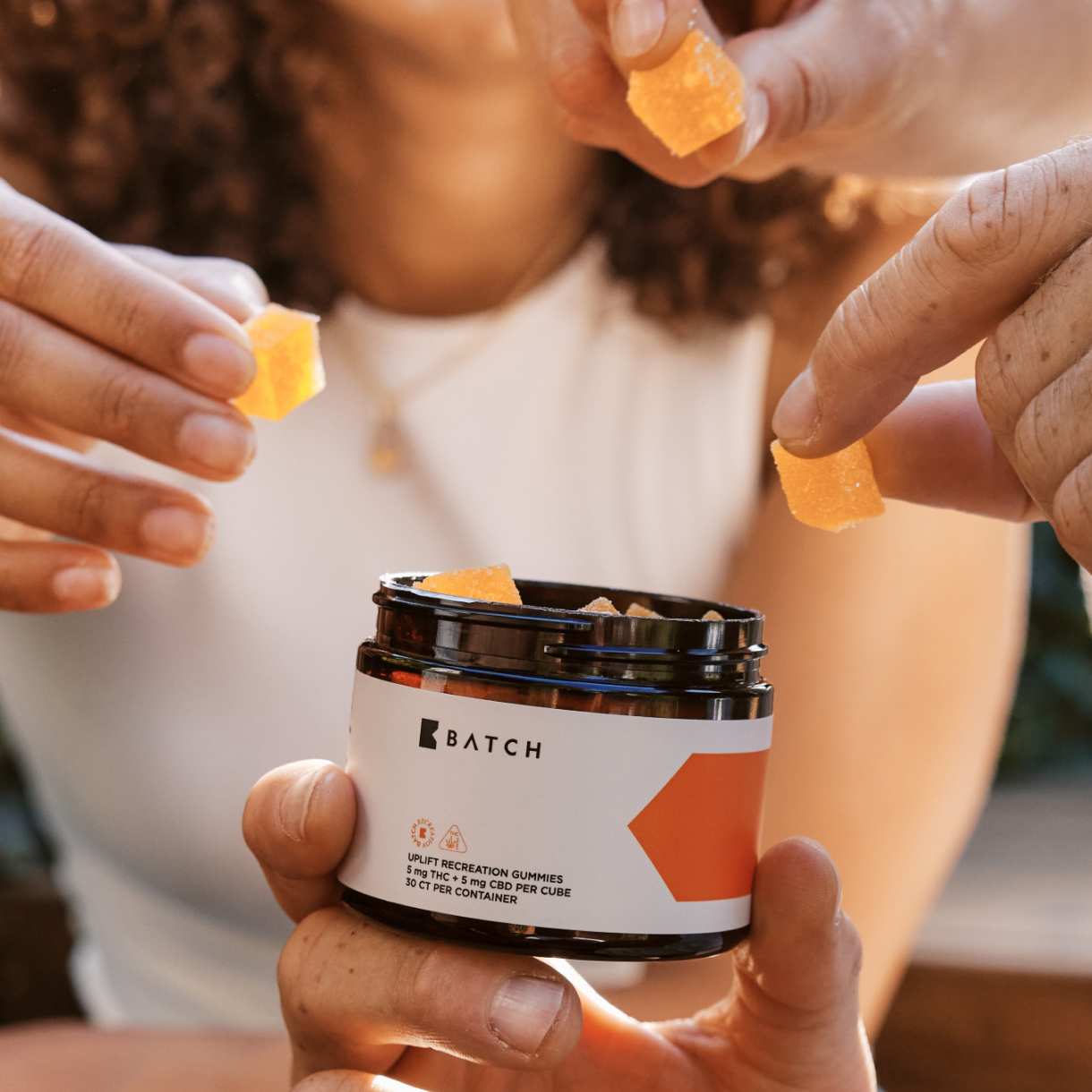 Close-up of several hands taking orange cube-shaped gummies from an open jar of Batch gummies.