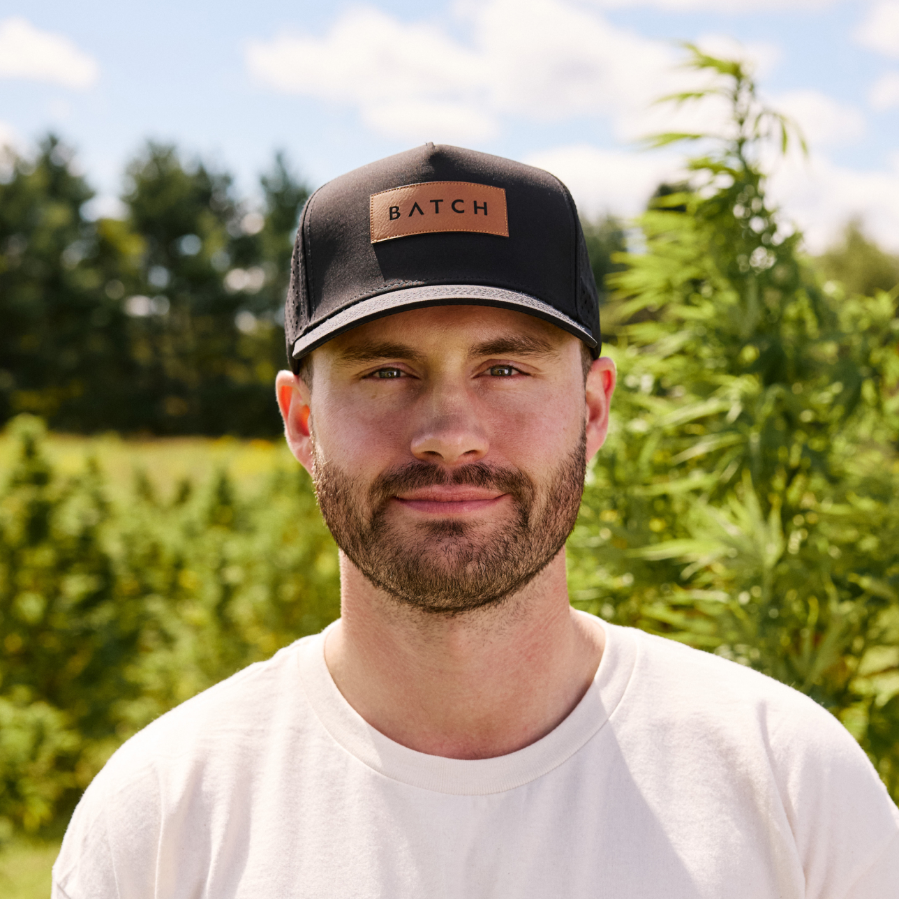 A man with a beard wearing a black 'BATCH' hat and white t-shirt, standing outside in a field.