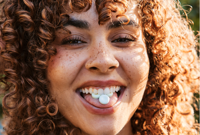 Person with curly hair smiling, holding a white candy on their tongue.