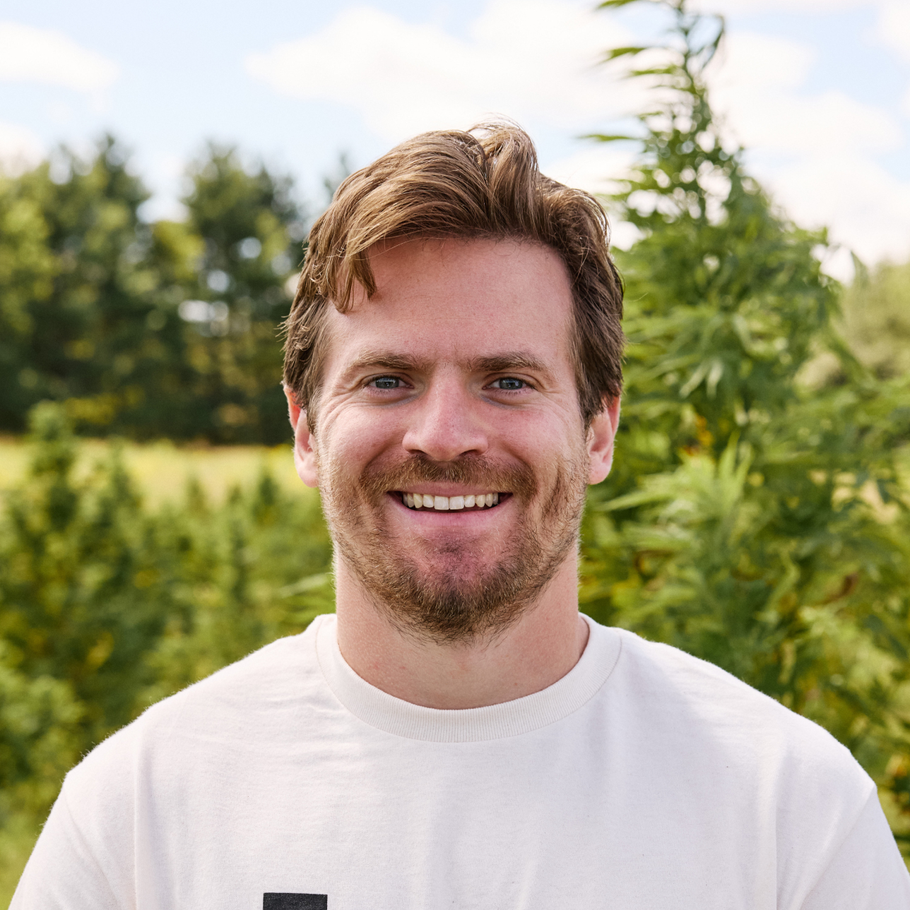 A smiling man with light brown hair and a beard in a white t-shirt, standing outside.