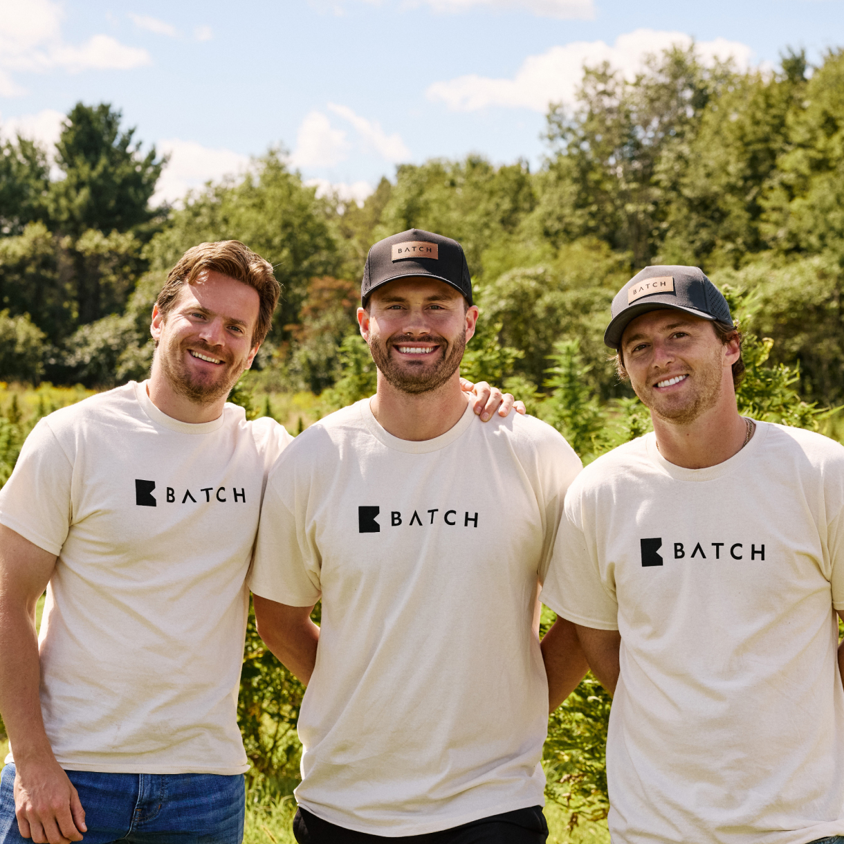 Three men wearing BATCH branded t-shirts and hats, smiling together in an outdoor field.