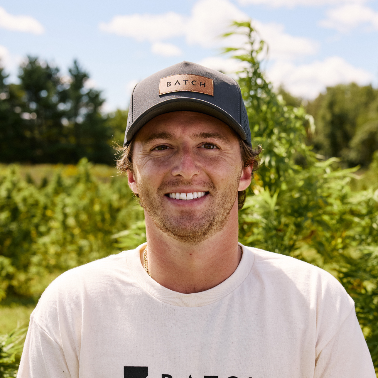 A smiling man wearing a grey 'BATCH' hat and a white t-shirt, standing in a field.