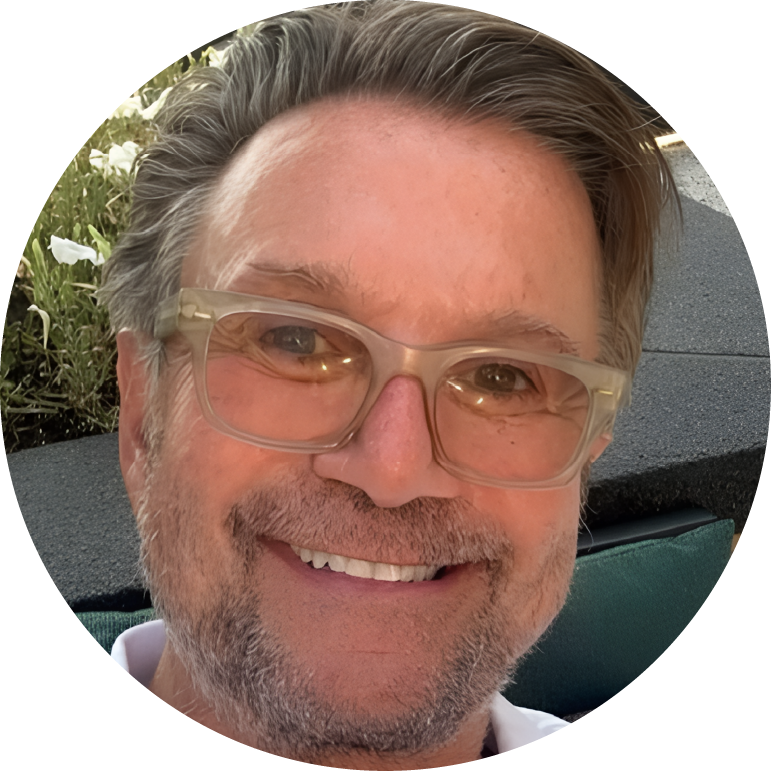 A person with glasses smiling, outdoors with flowers in the background.