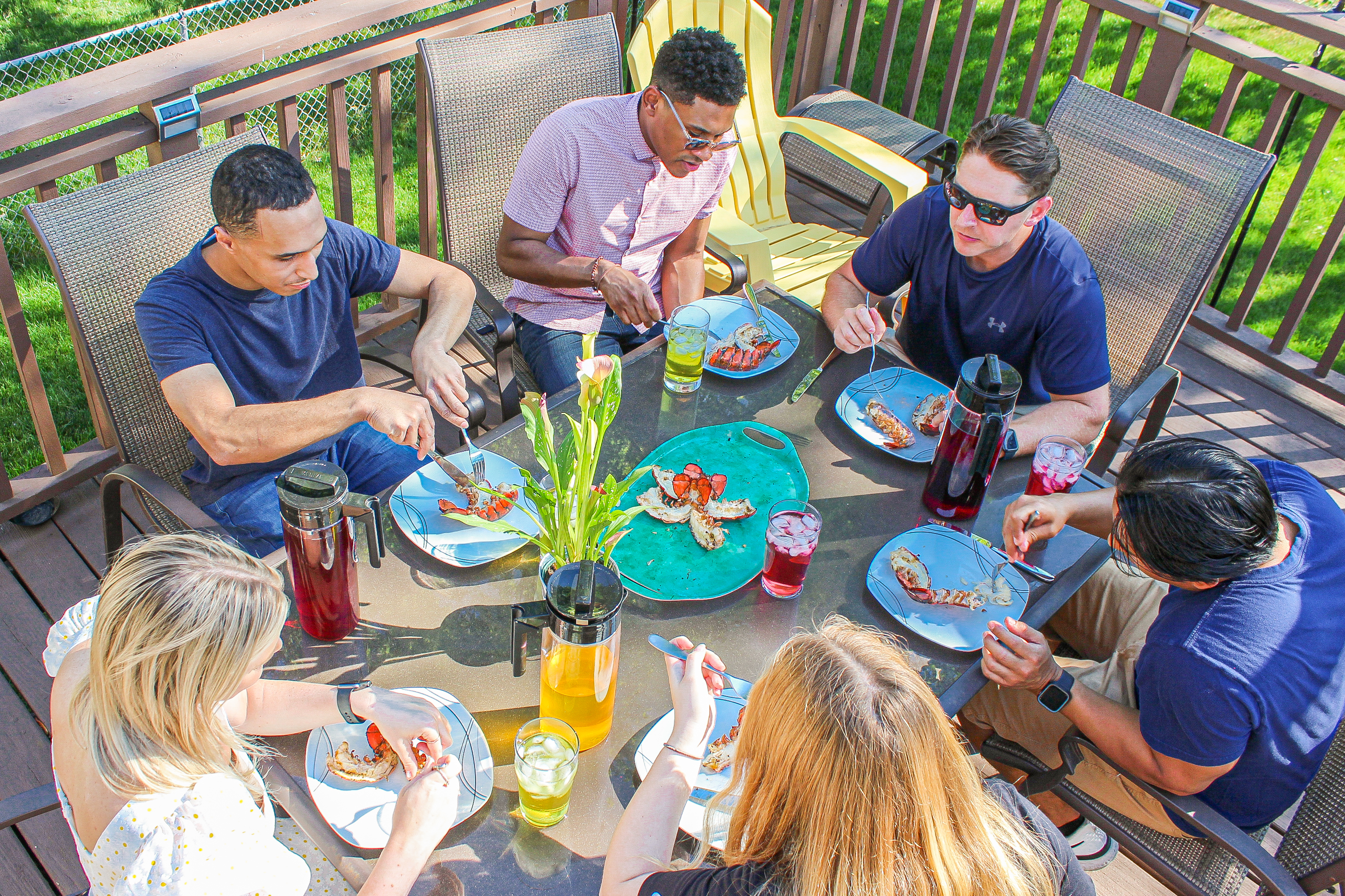 An overhead view of six people eating a lobster meal at a glass table on an outdoor deck.
