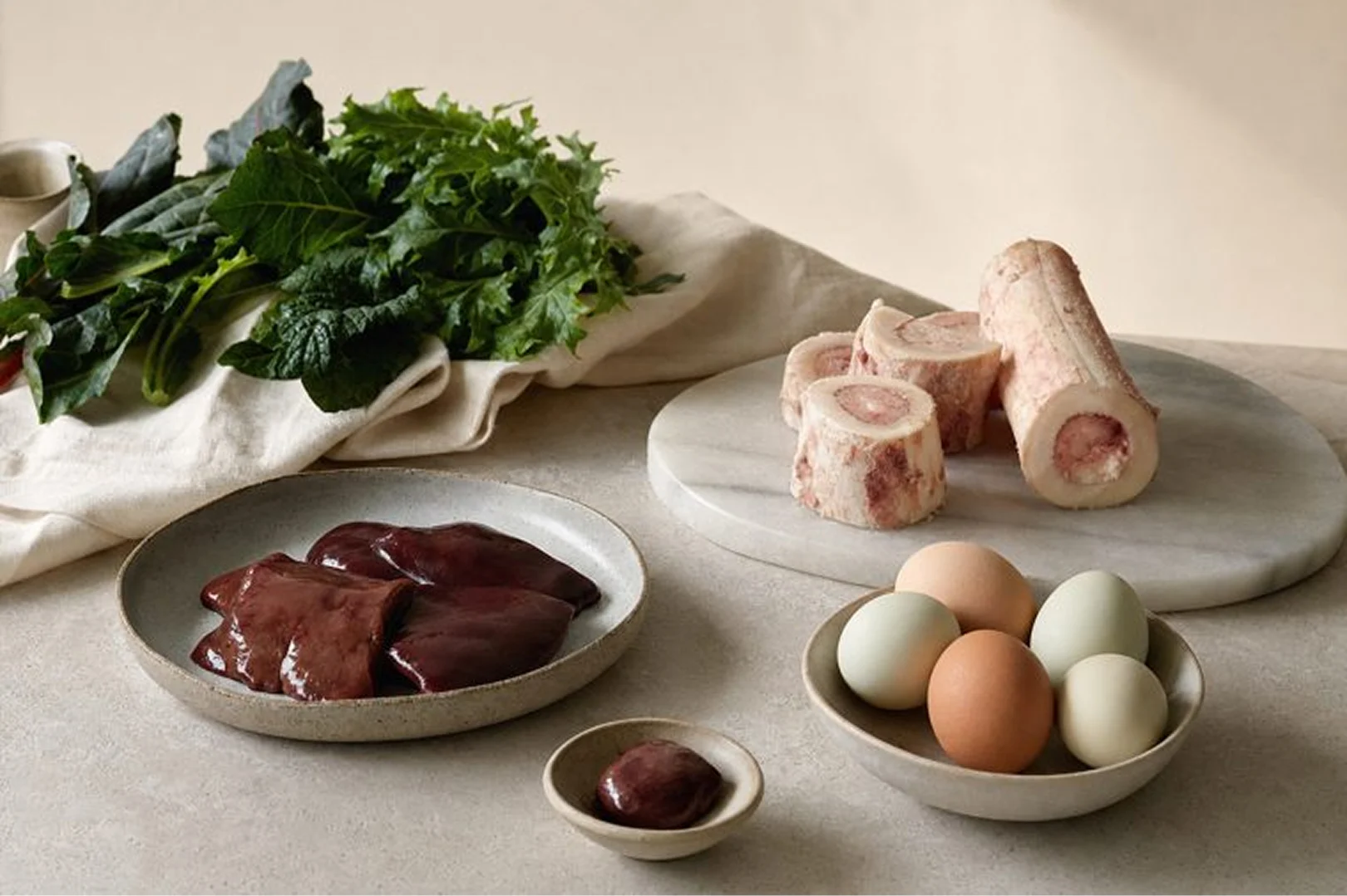 A tabletop arrangement of raw liver, marrow bones, a bowl of eggs, and a bunch of leafy greens.