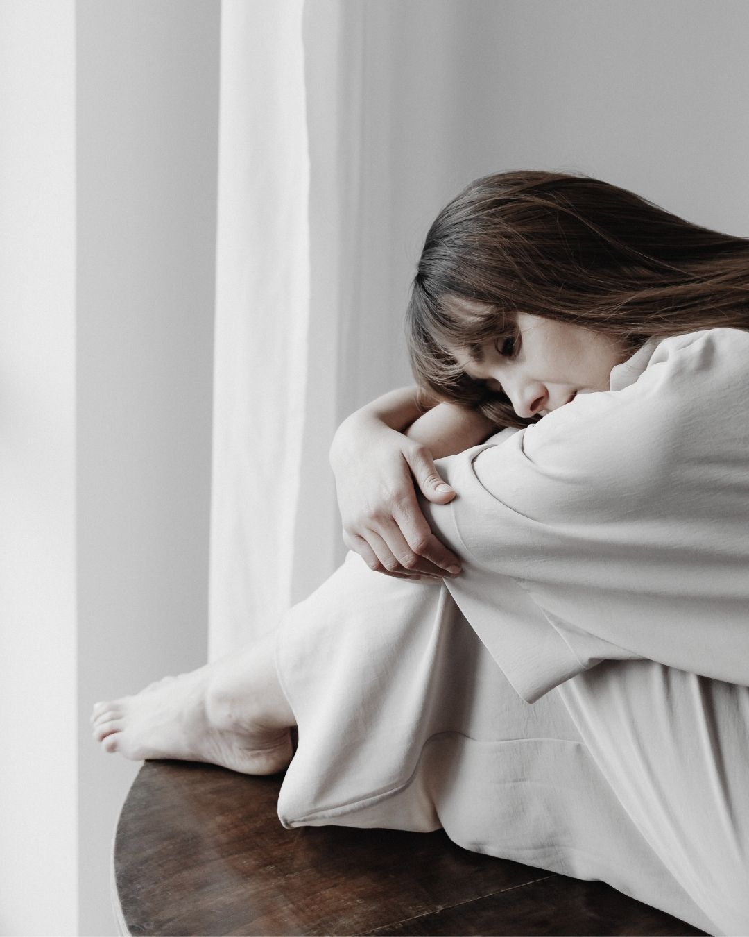 Person seated, resting head on knees, wearing a light-colored garment.