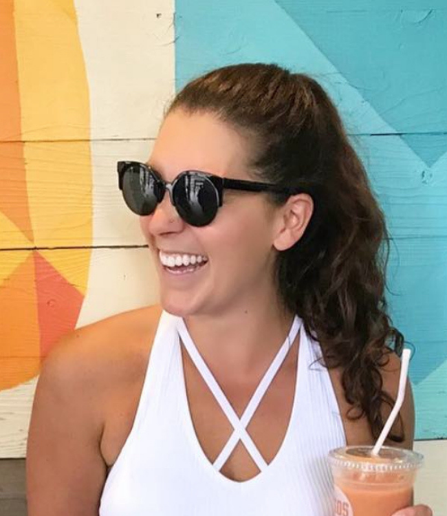 Person smiling with sunglasses, holding a drink against a colorful background.