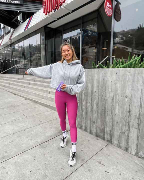 Person in pink leggings and grey hoodie smiling outside a building.