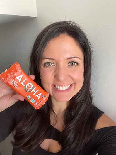 A person smiling and holding an Aloha protein bar in an orange wrapper.