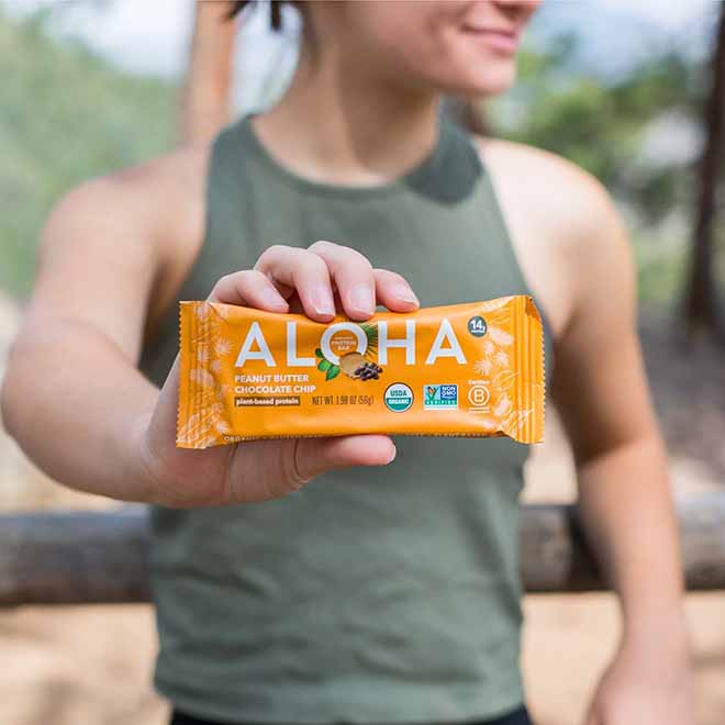 Person holding an Aloha peanut butter chocolate chip protein bar outdoors.