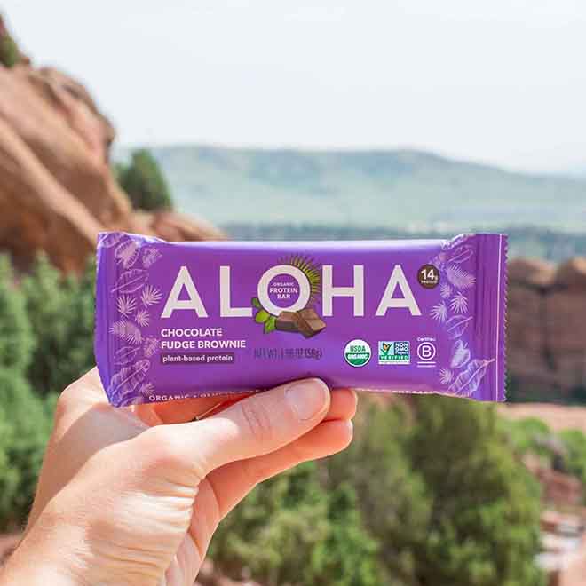 Hand holding ALOHA chocolate fudge brownie protein bar outdoors.