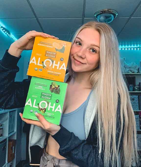 Person holding two boxes of Aloha bars, smiling in a room with blue lighting.