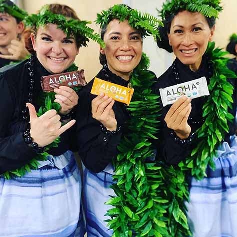 Three people in Hawaiian attire holding Aloha snack bars and smiling.