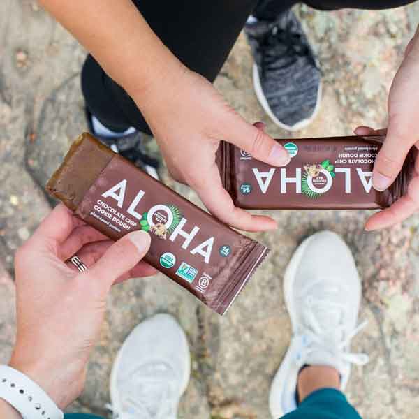 Two people holding Aloha chocolate chip cookie dough protein bars outdoors.