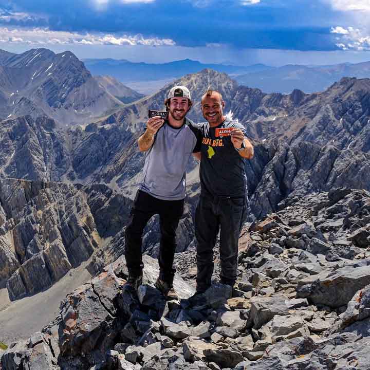 Two people smiling and holding cards, standing on a rocky mountain peak.