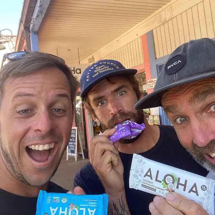 Three men holding Aloha snack bars, smiling in front of a store.