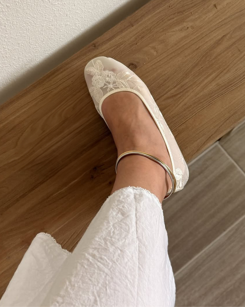 Foot wearing a white lace shoe and a gold anklet on a wooden surface.