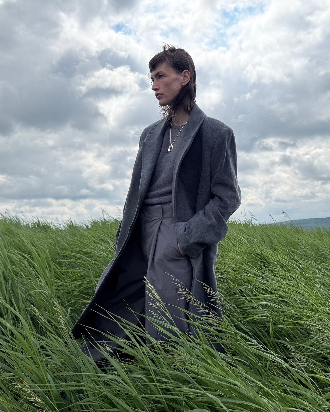Person in a gray coat standing in a windy grassy field under a cloudy sky.