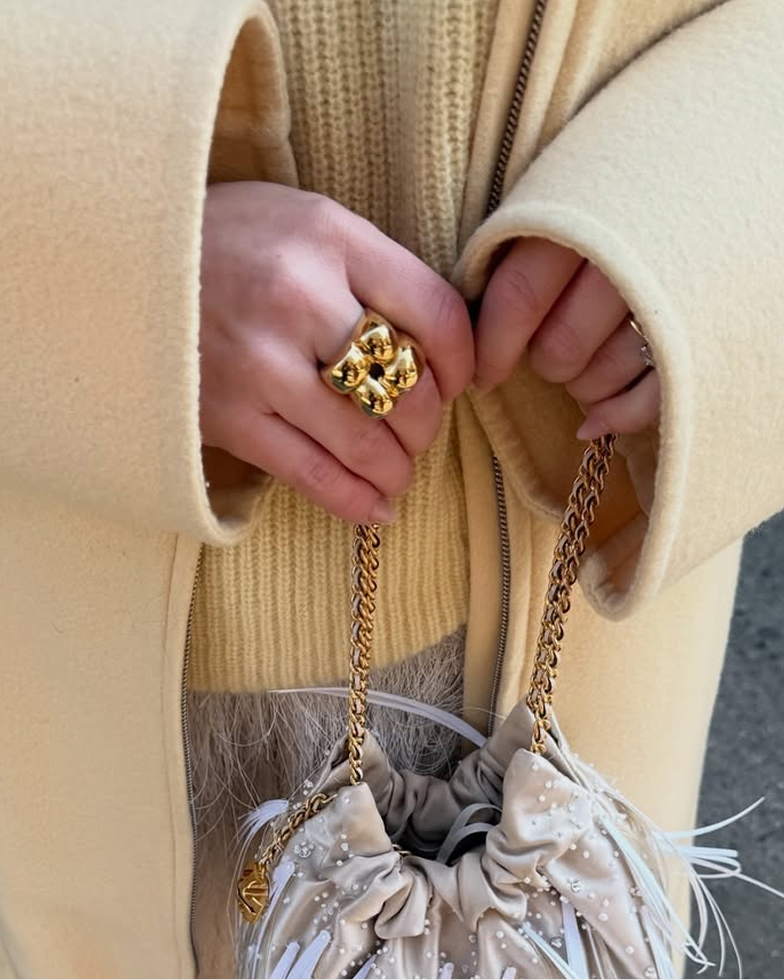 A close-up of a person's hands wearing a chunky gold ring and holding a feathered, beaded bag.