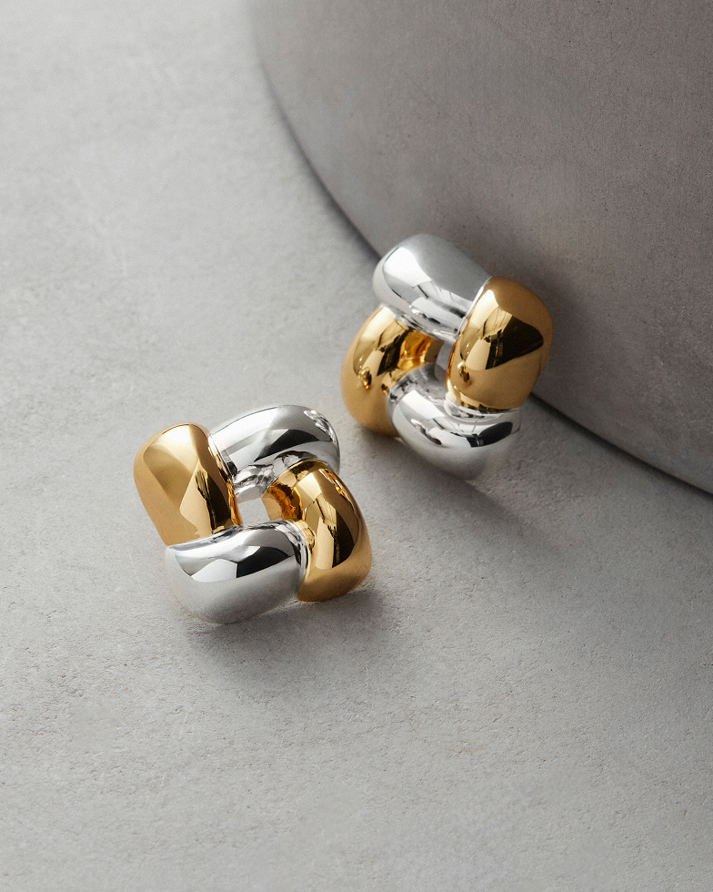 A pair of chunky, interlocking earrings in polished gold and silver tones on a gray surface.